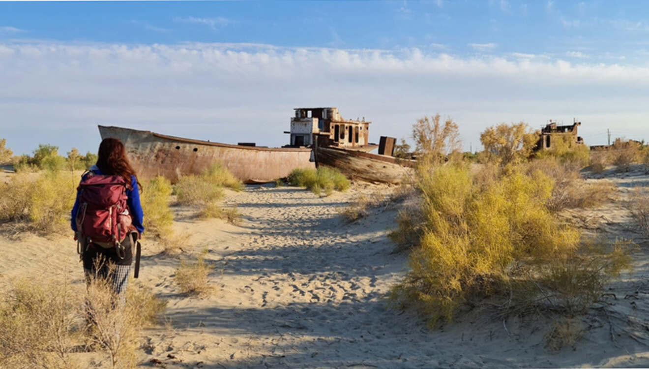 A person with a backpack walks toward a large abandoned ship stranded in a desert landscape with sparse vegetation and blue sky.