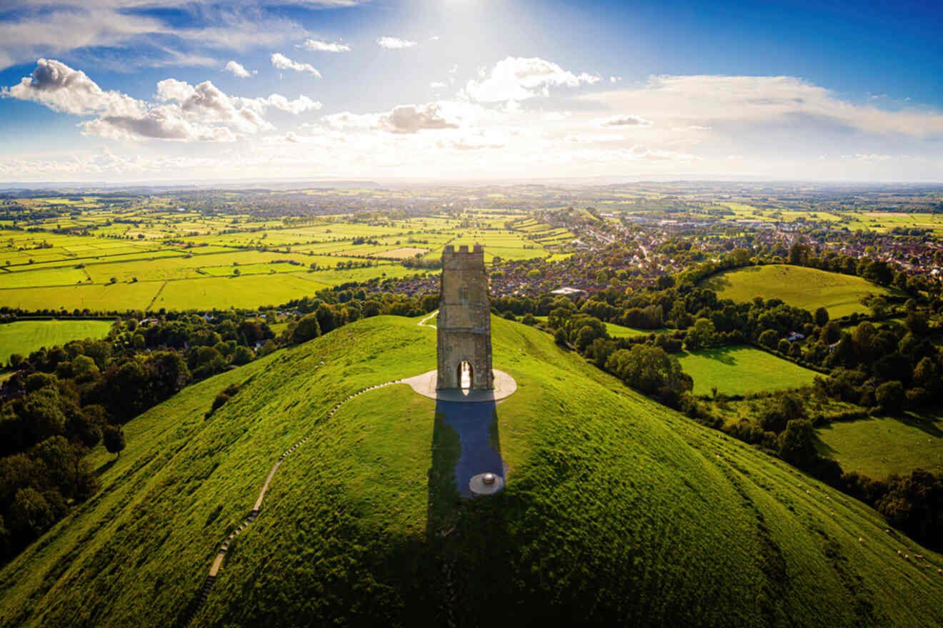 An aerial view of Glastonbury Tor with its iconic tower on a hilltop surrounded by lush green fields.