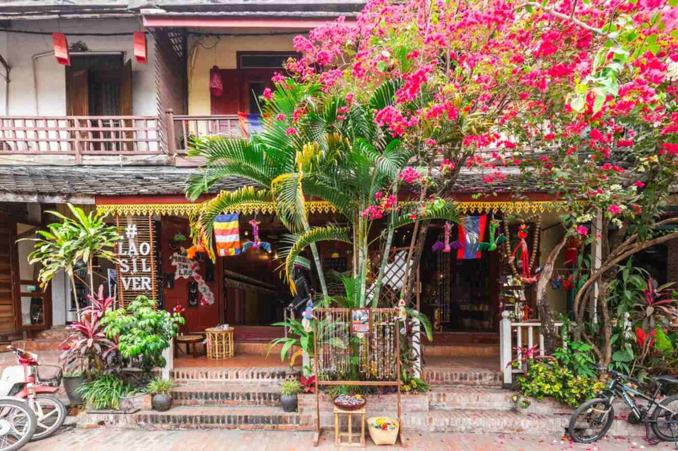 A storefront adorned with colorful fabrics, plants, and flowers, displaying a sign that reads "Lao Silver." Bicycles are parked in front and various items are hung under the roofed area.