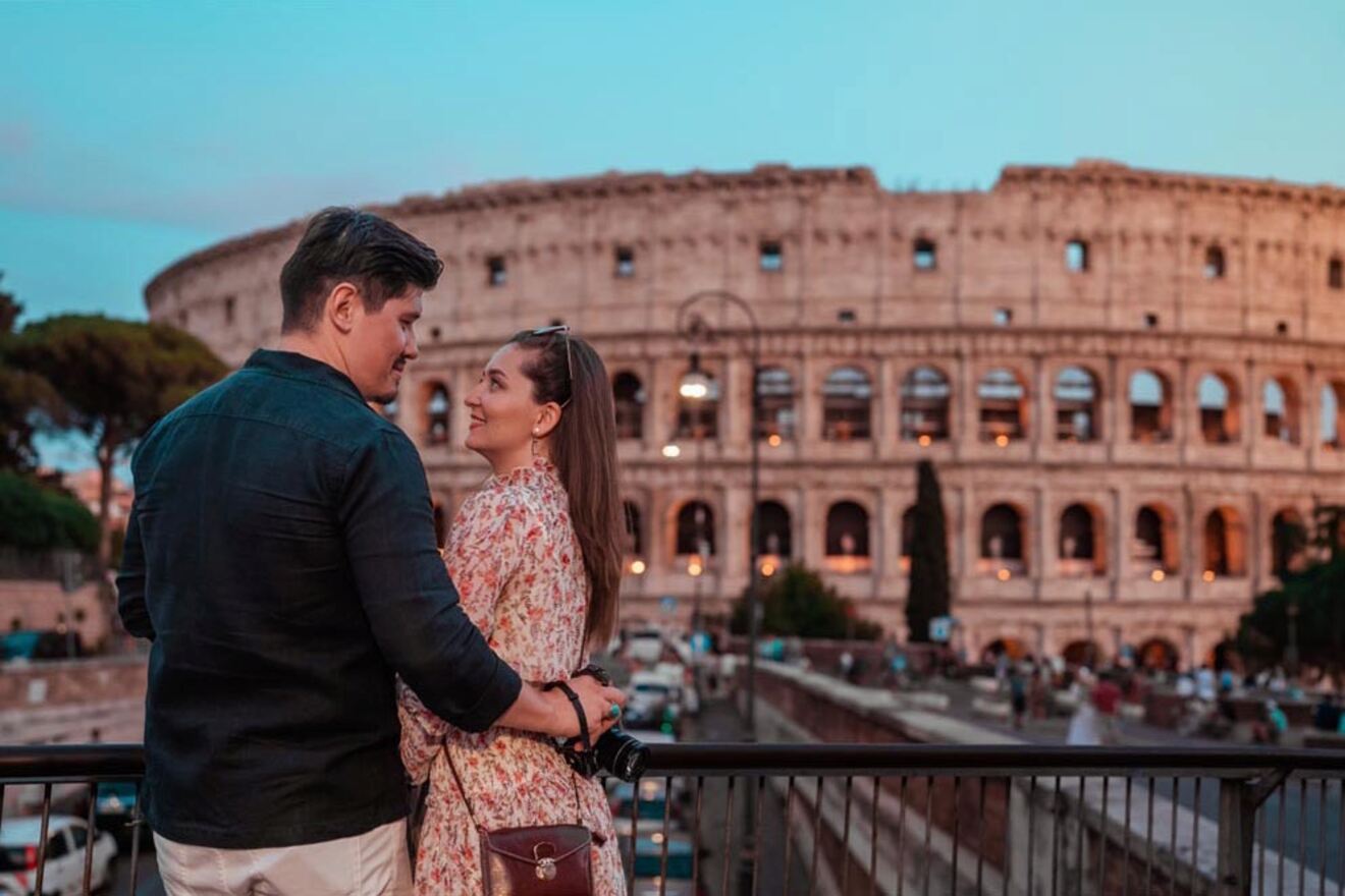 A couple stands on a balcony, facing each other, with the Colosseum in Rome in the background at sunset. The woman holds a camera, and both are casually dressed.