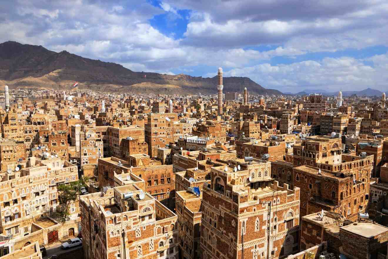 Aerial view of a densely built city with traditional brown buildings, numerous minarets, and distant mountains under a partly cloudy sky.