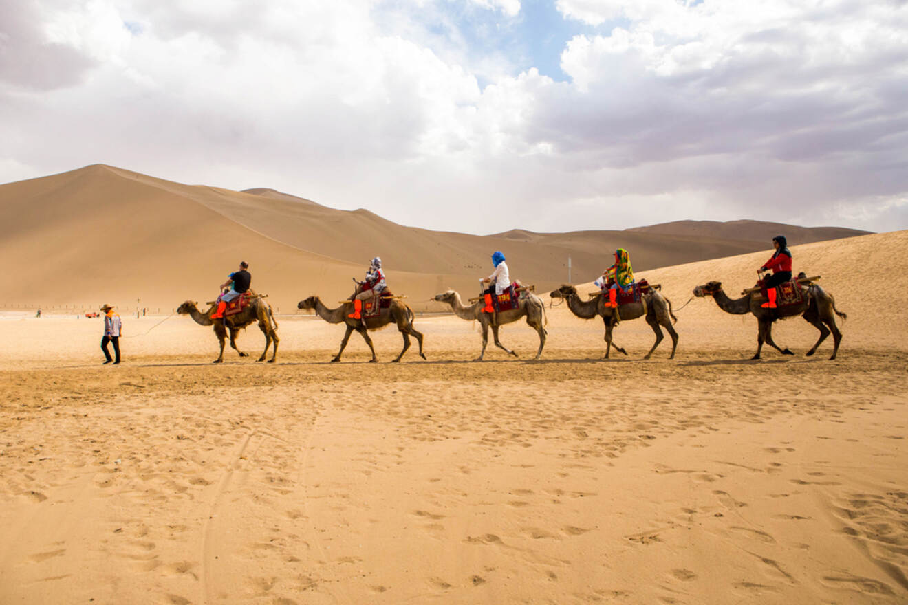 A line of tourists riding camels through the vast Gobi Desert, with sand dunes stretching out into the horizon.