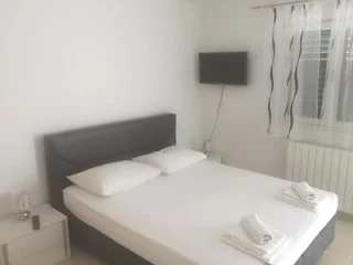 A minimalist bedroom with a double bed, two pillows, and neatly folded towels. A small nightstand is beside the bed, and a flat-screen TV is mounted on the wall. A window with white curtains is visible.