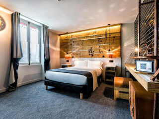 A modern hotel room with a queen-sized bed, wooden headboard with floral design, bedside tables, desk, chair, and large windows with curtains.