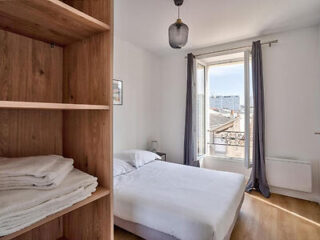 A bright bedroom with a double bed, wooden shelving unit stocked with folded towels, a hanging light fixture, and a window with gray curtains, partially opened to reveal a cityscape view.