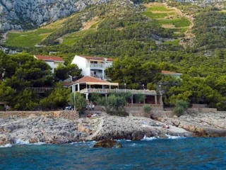 Coastal vacation homes surrounded by trees at the base of a rocky hillside, with a rocky shoreline and blue sea in the foreground.