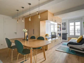 A modern living area features a wooden dining table with green chairs, pendant lighting, a modular wooden storage unit with a built-in seat, and a gray sofa next to a window with natural light.