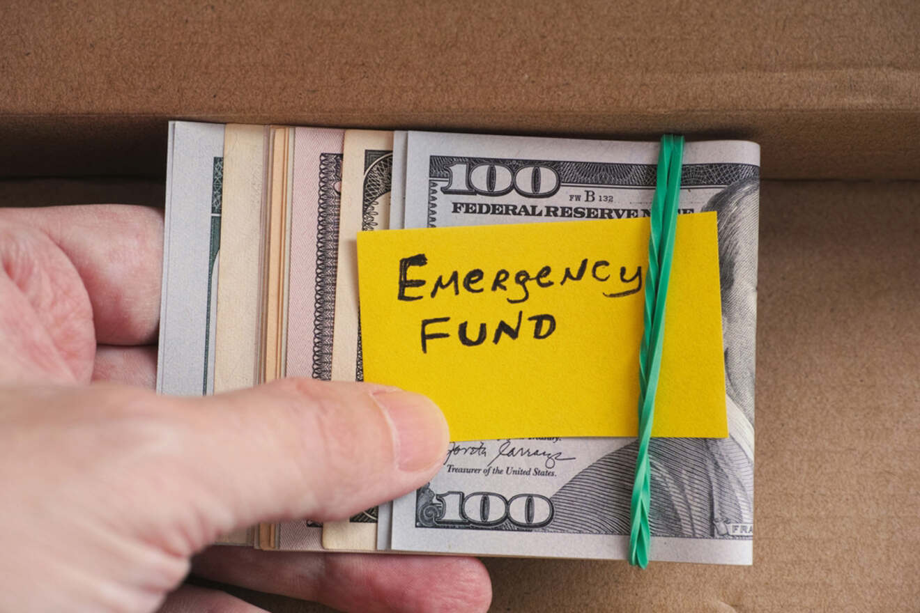 A bundle of 100-dollar bills, secured with a green rubber band, labeled "Emergency Fund" on a yellow sticky note being held by a hand inside a cardboard box.