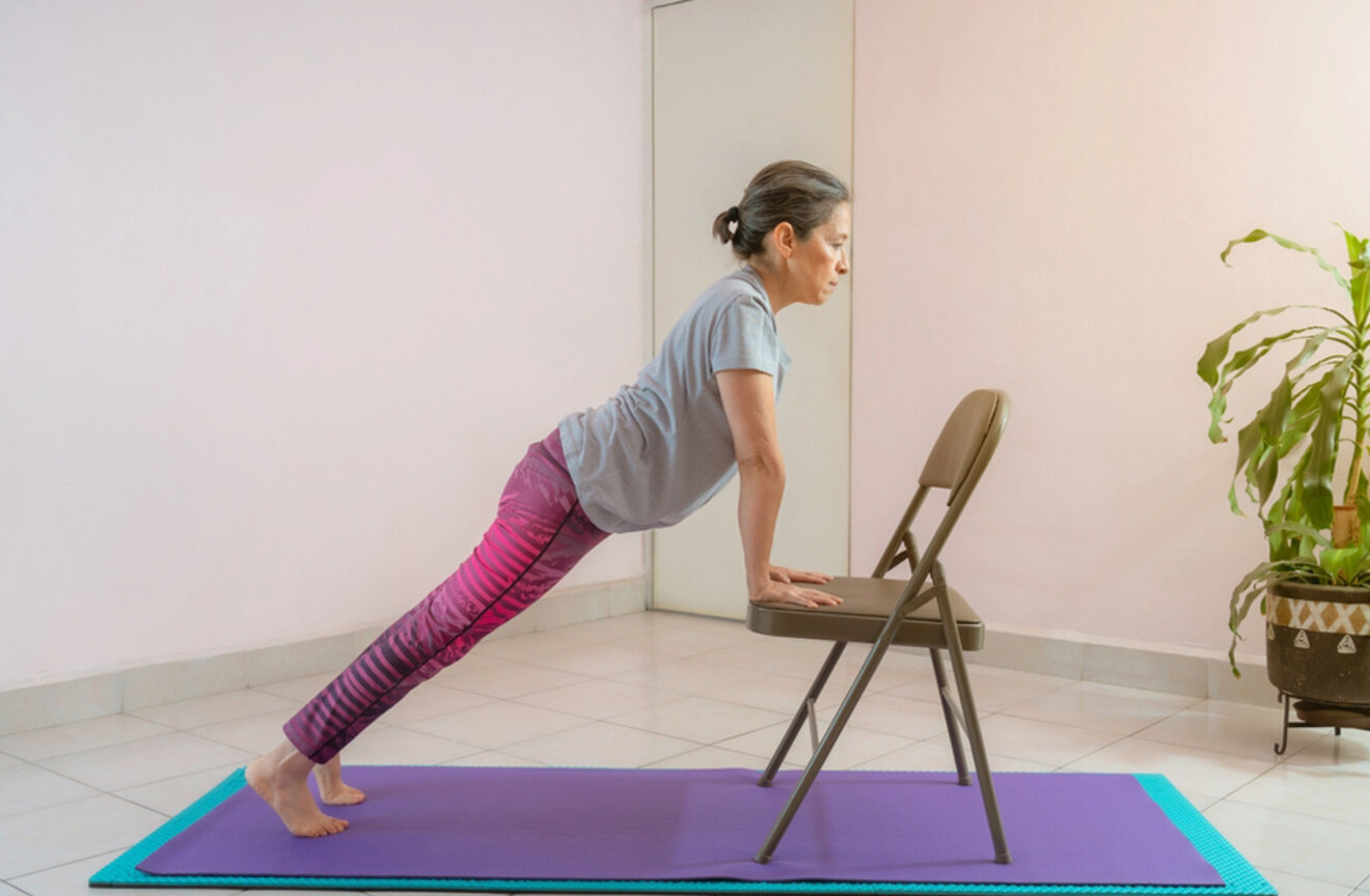 An older woman practicing a modified plank pose using a chair for support on a purple mat in a minimalistic room.