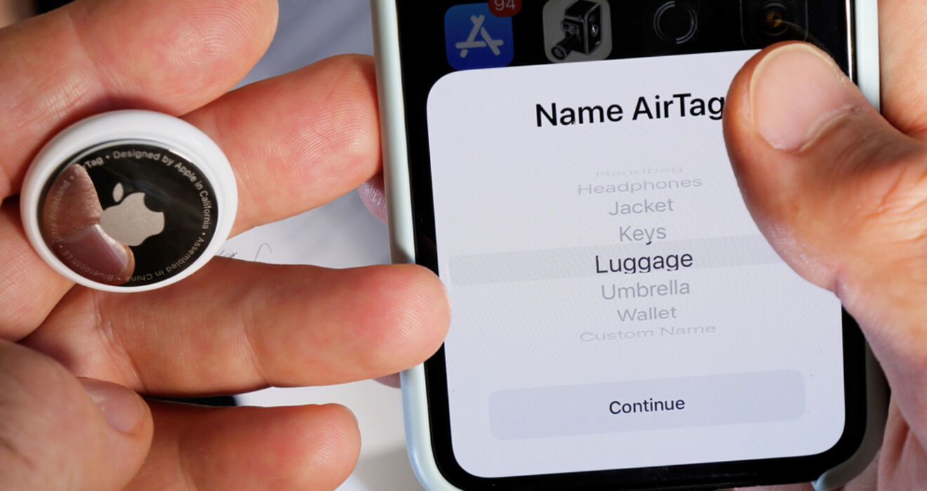 A person holding an Apple AirTag next to a smartphone screen displaying setup options for naming the AirTag, including options like "Headphones," "Jacket," "Keys," and "Luggage.