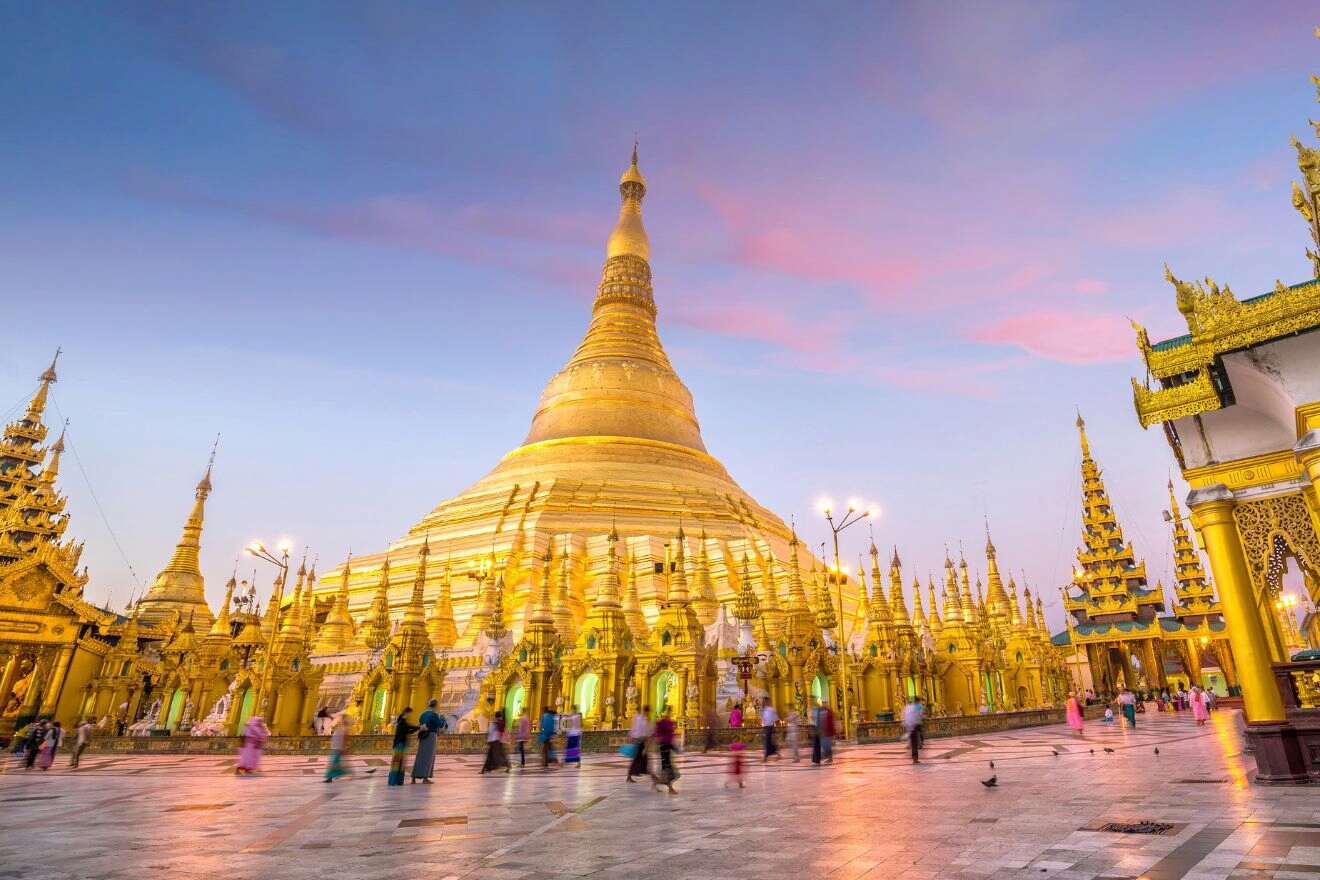 Golden pagoda with a tall spire at sunset, surrounded by smaller structures and visitors walking around. Sky shows blue and pink hues.