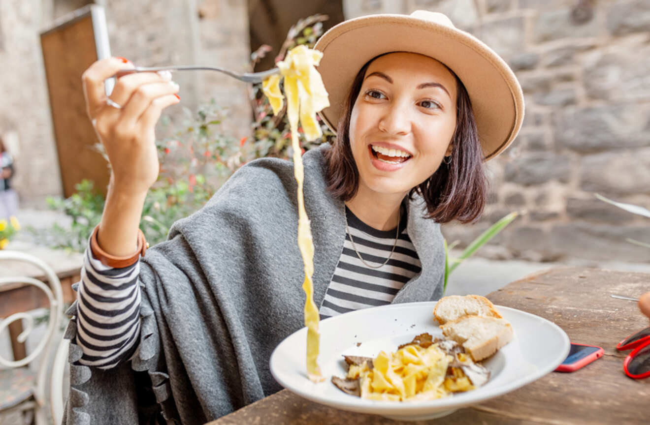 Woman in a hat smiling while holding up a forkful of pasta at an outdoor restaurant in Rome, Italy.