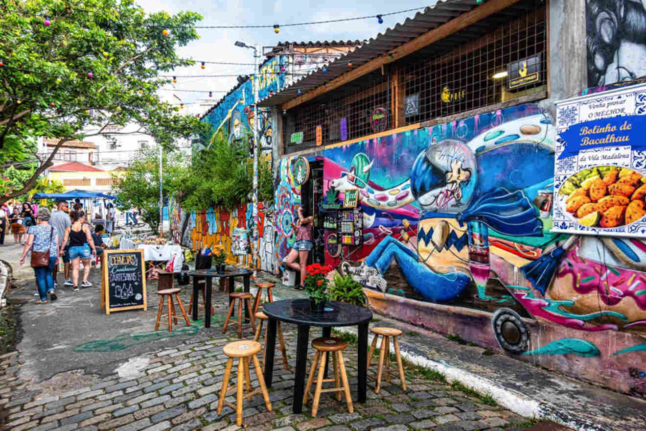 A lively alleyway features vibrant street art and colorful murals on the walls. Outdoor seating with wooden stools and tables is available, and a few people are seen walking and exploring the area.