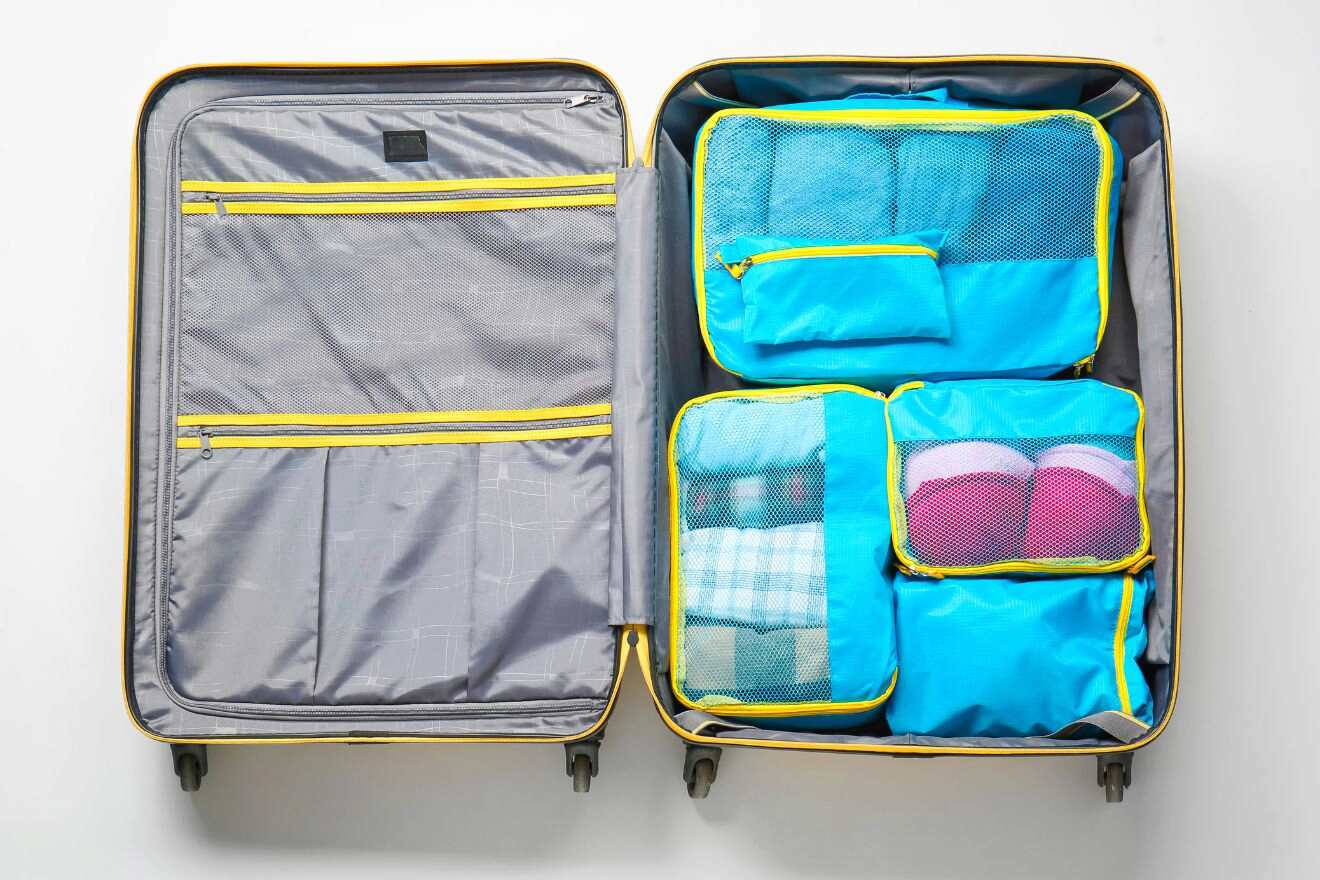An open suitcase contains blue packing cubes filled with folded clothes. The right side has four packing cubes, and the left side has zippered compartments. The suitcase has a yellow interior lining.