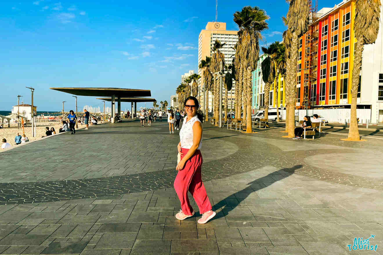 Darija Stojanovic, la autora de las publicaciones de misstourist, con pantalones rosas y gafas de sol, camina por un paseo marítimo pavimentado con edificios coloridos y palmeras en el fondo.