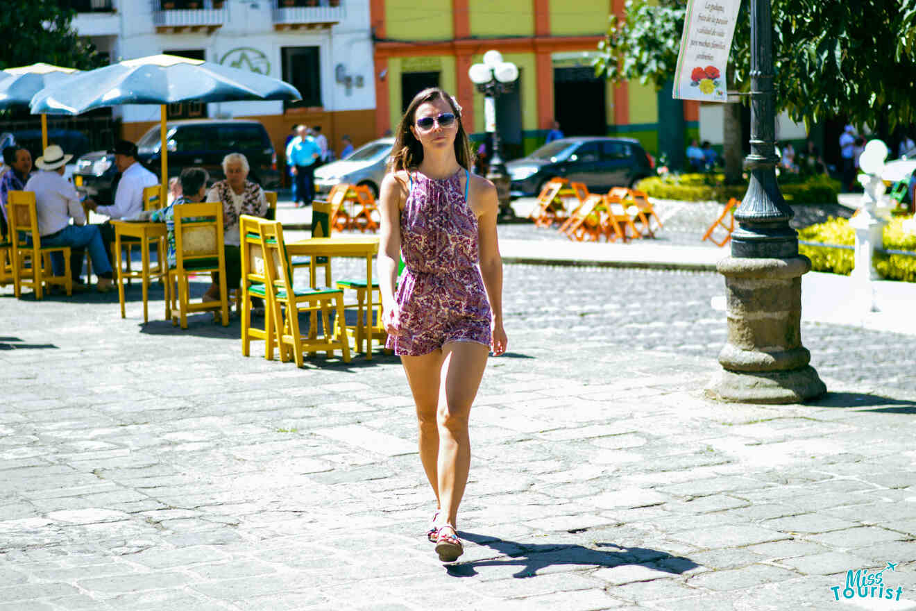 Yulia, la fundadora de este sitio web, con un mono estampado, camina por una zona pavimentada al aire libre con mesas y sillas. Al fondo, hay gente sentada bajo una gran sombrilla. Se ven un cartel y edificios.