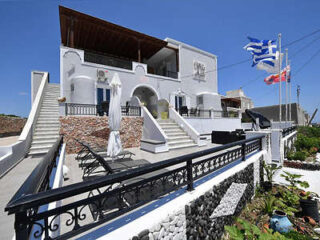 A two-story house with a modern white facade featuring multiple balconies, stone accents, and several flags flying on tall poles in the front yard. Steps lead up to the house from the paved pathway.