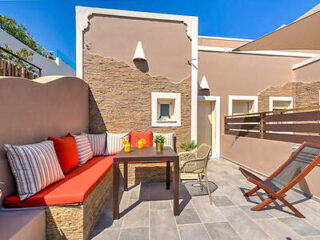 A sunny outdoor patio with a cushioned bench, a wooden table with drinks and plants, two chairs, and decorative stone walls.