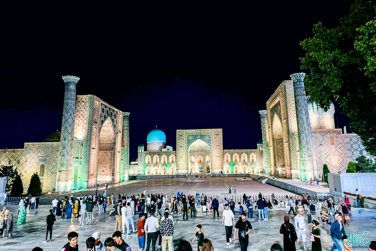 A night view of Registan Square in Samarkand, Uzbekistan, showcasing its illuminated madrassahs with a crowd of people walking and taking photos in the foreground.