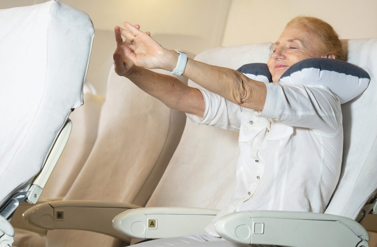 An elderly woman stretches her arms while sitting on an airplane, using a neck pillow for support.