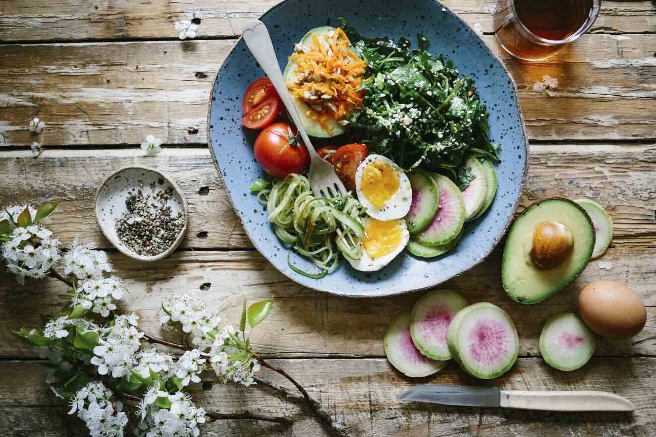 A bowl with avocado, hard-boiled egg, tomatoes, zucchini noodles, and leafy greens on a rustic wooden table with a knife, fresh avocado, radishes, and a small plate of pepper. White flowers are visible.