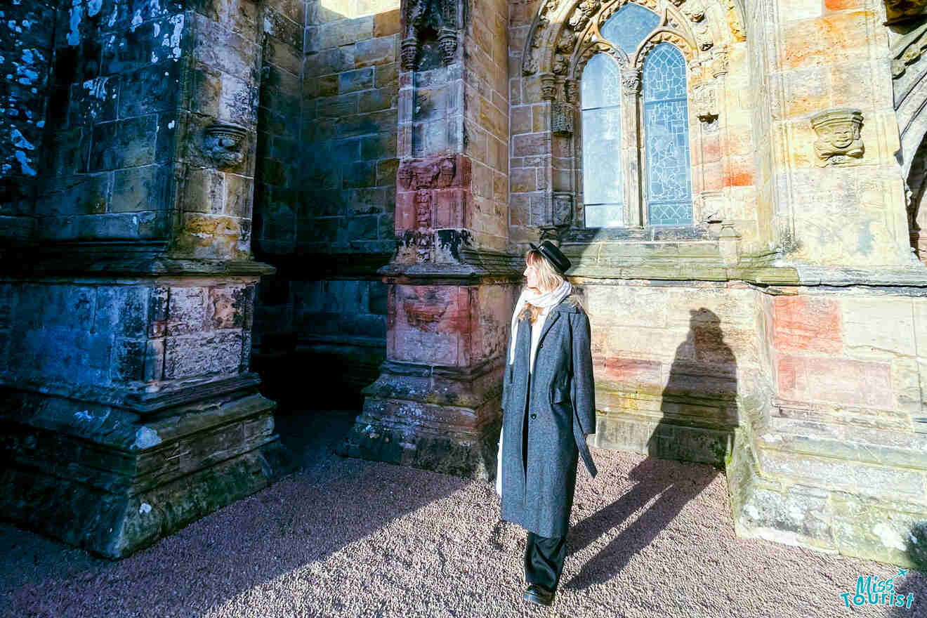 Victoria Lupascu, the author of the posts or misstourist, wearing a long coat and hat stands in front of a sunlit, weathered stone building with arched windows.