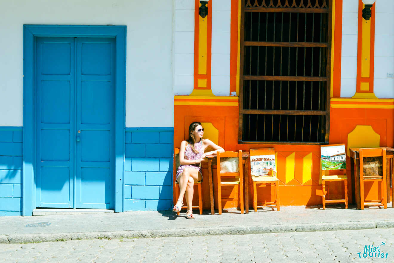 Yulia, la fundadora de este sitio web, está sentada en una silla de madera afuera de un edificio colorido con paredes azules, naranjas y blancas, junto a una puerta azul y varias sillas que exhiben pinturas.