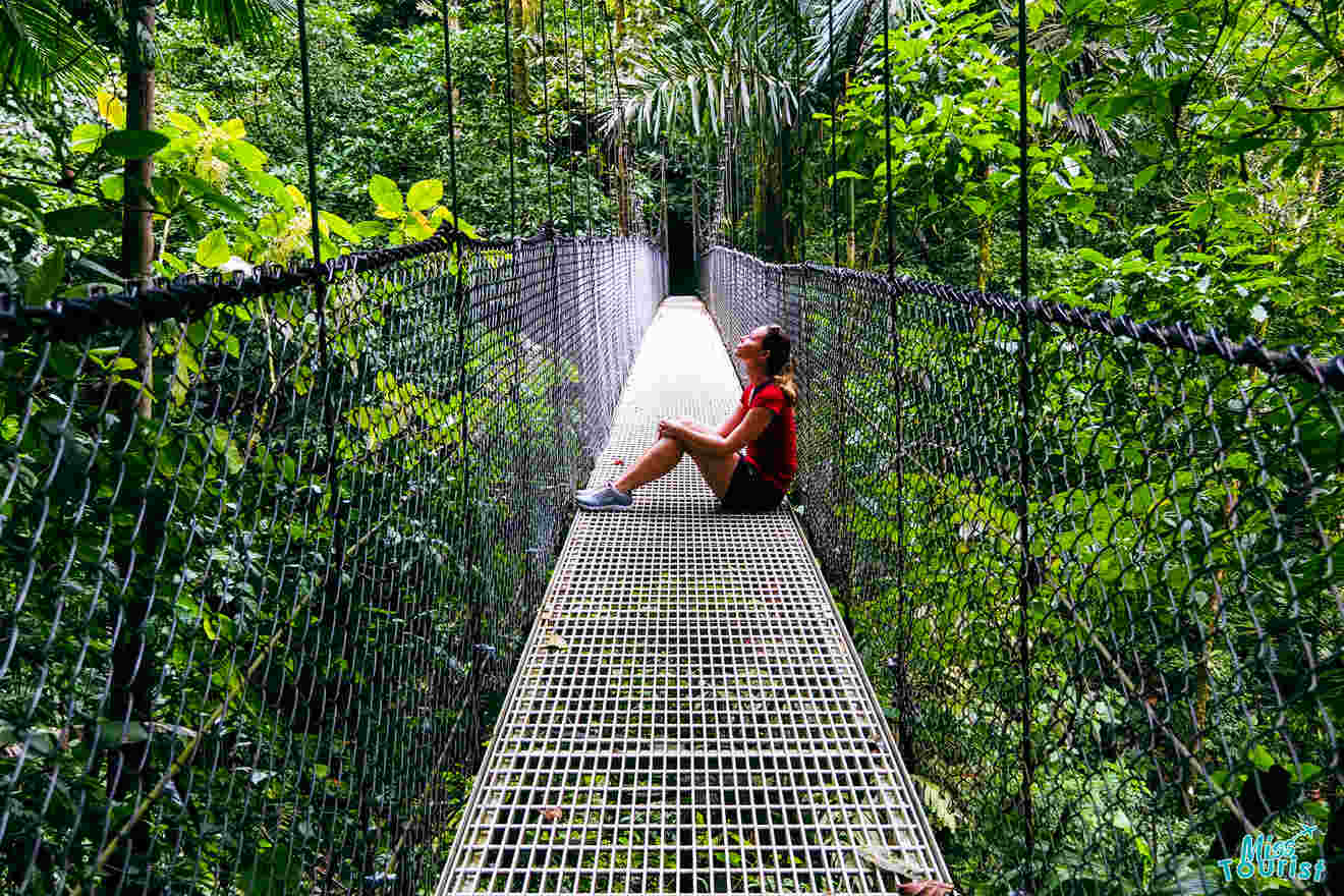 Yulia, la fundadora de este sitio web, se sienta con las piernas cruzadas en un estrecho puente colgante rodeado de exuberante follaje verde.
