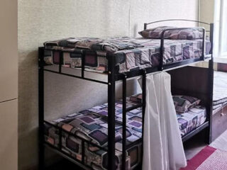A metal bunk bed with geometric-patterned bedding is placed against a wall; a white curtain hangs beside the bottom bunk near a window.