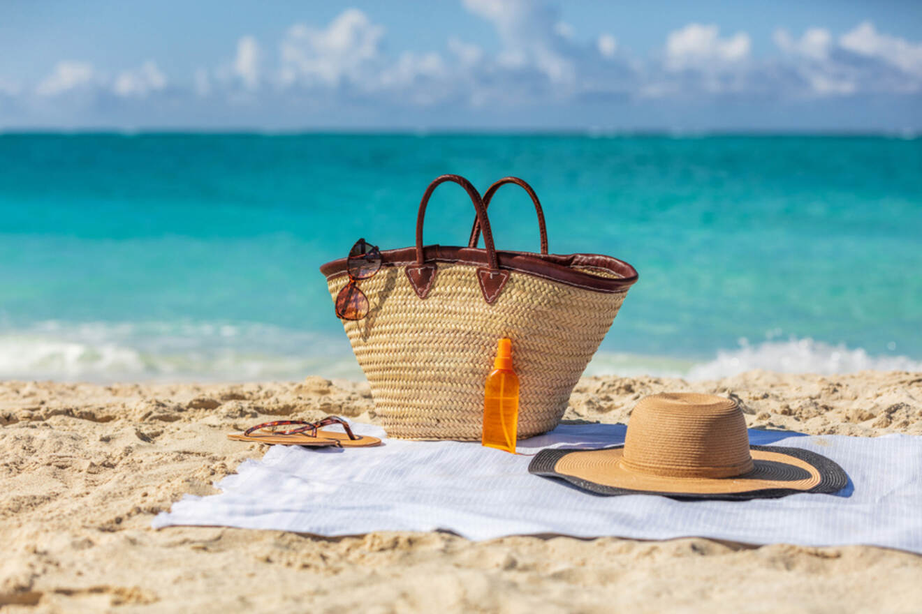 A straw beach bag, sunglasses, sunscreen, and a sunhat are placed on a white towel on a sandy beach with turquoise water in the background.