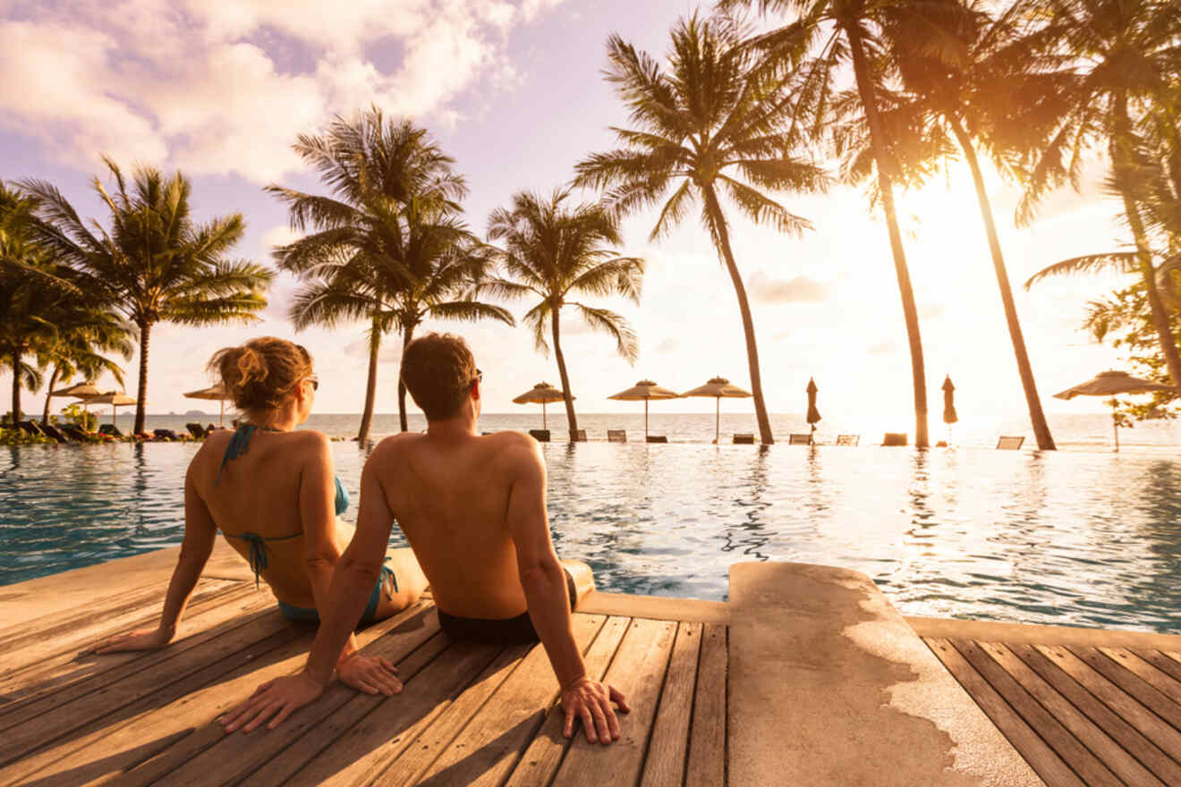 Two people sit at the edge of a pool overlooking a beach with palm trees and sun umbrellas at sunset.