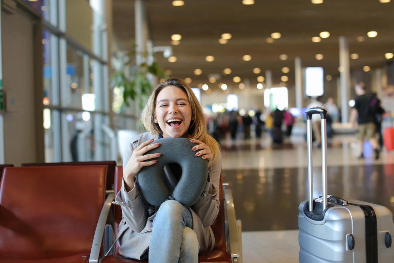 A woman sits in an airport lounge, holding a neck pillow and smiling. A suitcase is next to her, and other travelers can be seen in the background.