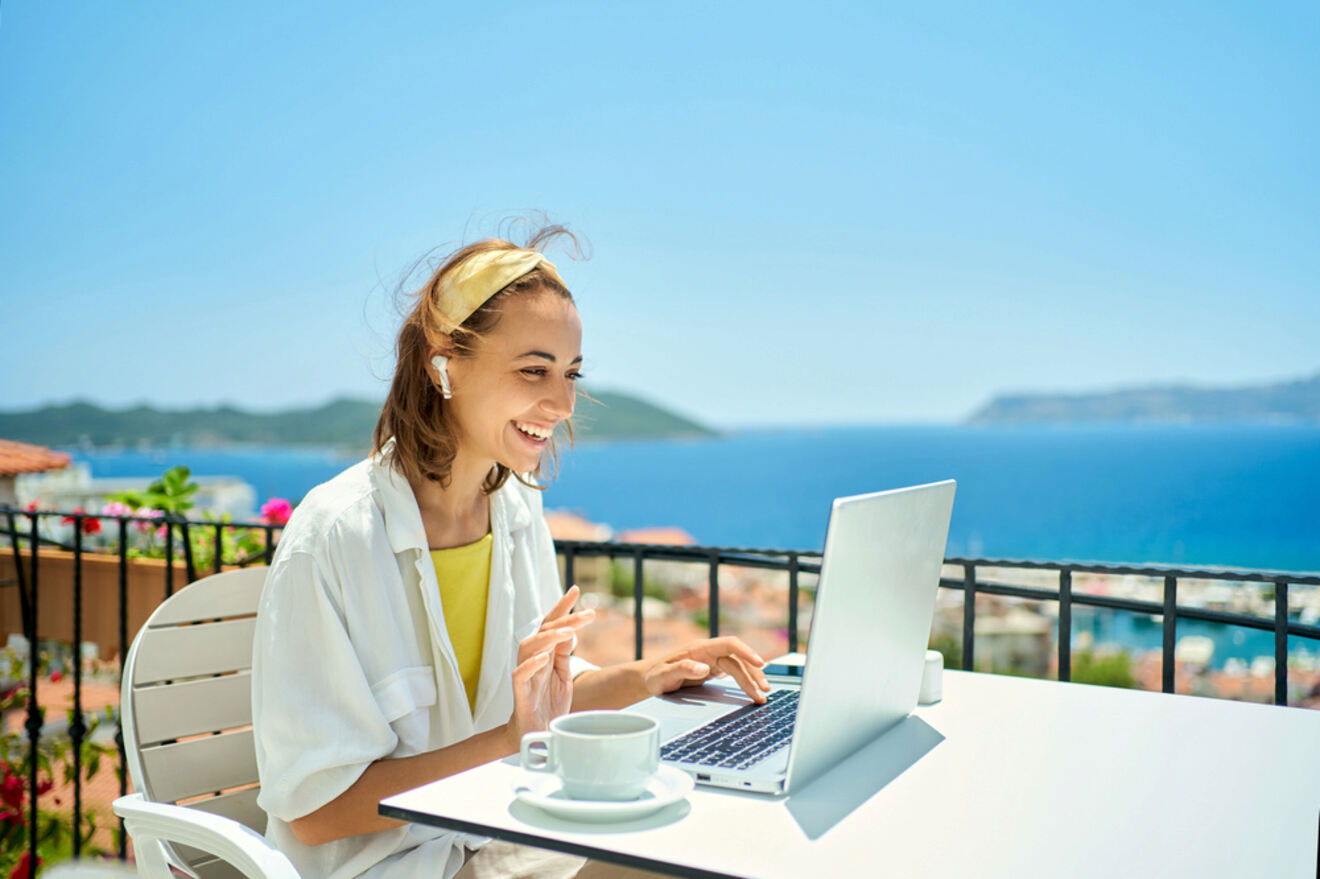 A woman sits at an outdoor table with a laptop and a cup of coffee, smiling, with a scenic coastal view in the background.