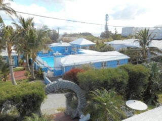 View of a residential area with blue houses, palm trees, and a stone arch in front. Power lines cross the scene, and the sky is partly cloudy.