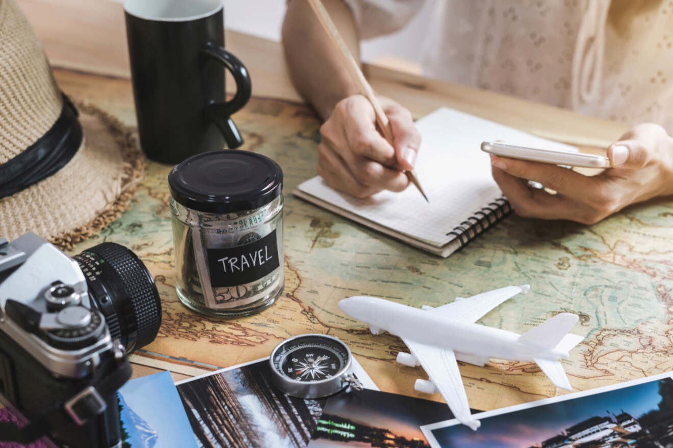 A person is planning a trip, writing in a notebook with travel items such as a map, camera, toy airplane, compass, and jar labeled 