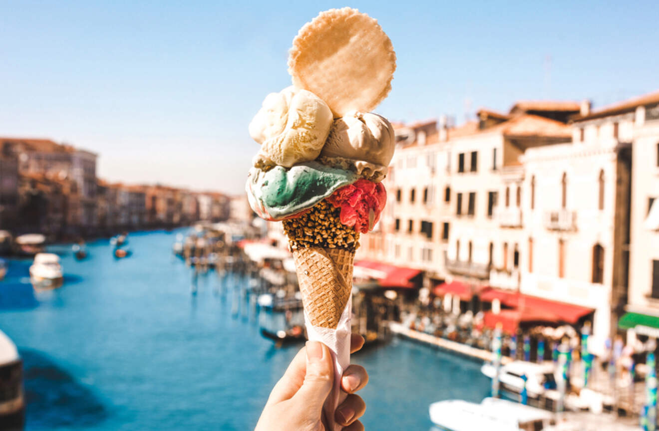 Hand holding a colorful gelato cone with a canal and buildings in the background in Venice, Italy