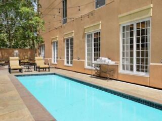 Narrow outdoor pool beside a beige building with several large windows, lounge chairs, a table, and string lights overhead.