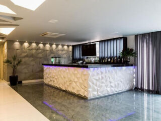 A modern bar with a textured white counter, backlit with blue light, bottles on shelves, a TV above, and geometric wall panels, in a bright room with large windows and gray curtains.