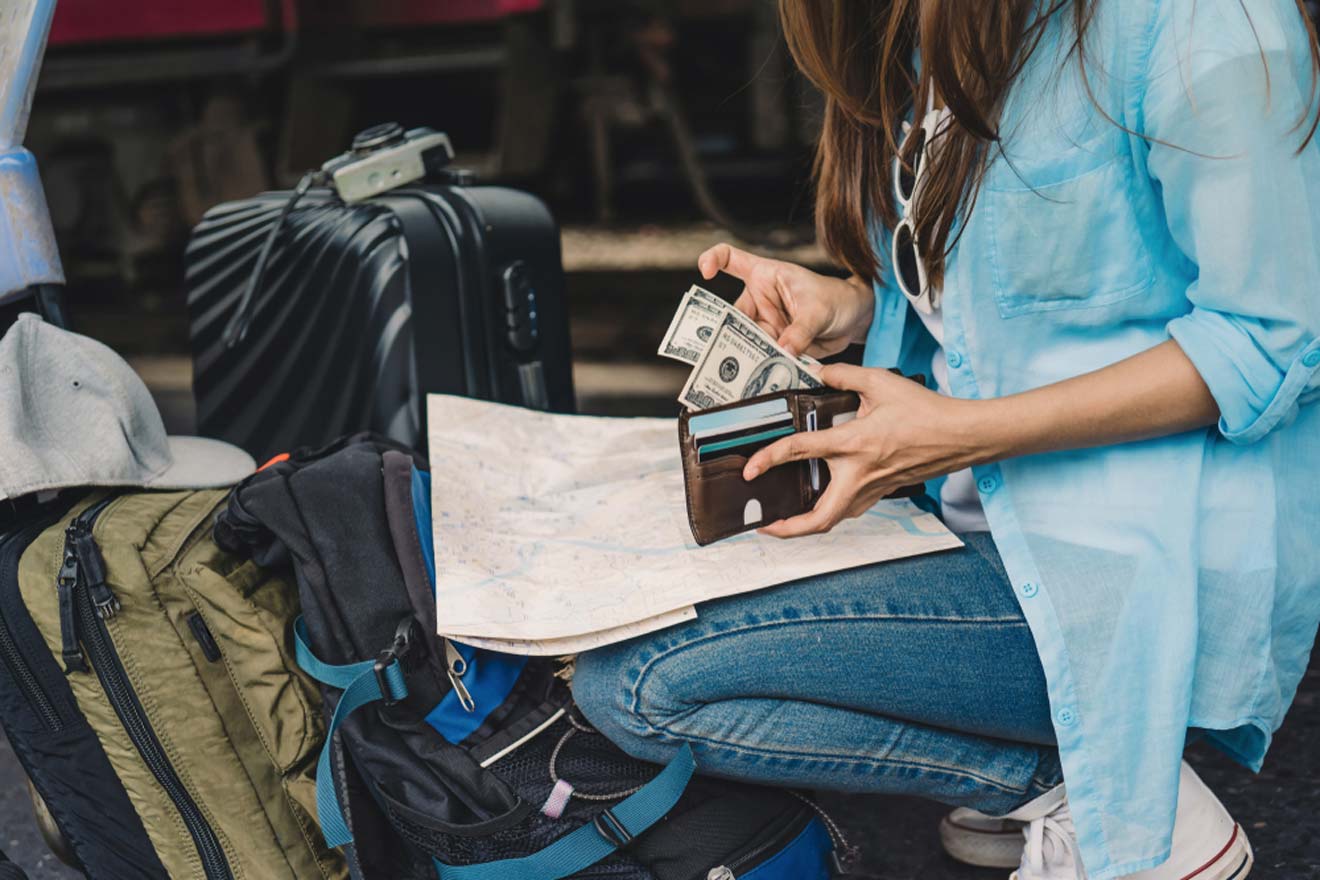 A woman in a blue shirt is holding dollar bills and a wallet, sitting near travel luggage and a map.