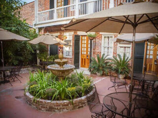 Brick courtyard with a central fountain, surrounded by patio tables, chairs, umbrellas, potted plants, and a two-story building with wooden doors and balconies in the background.