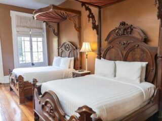 Two wooden canopy beds with white bedding in a room with brown walls, wooden floor, a lamp, and a window with white blinds.