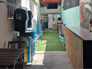 Outdoor seating area with colorful chairs, barrels, a wooden table, and T-shirts hanging on display. There is a counter along the right and a fridge in the back.