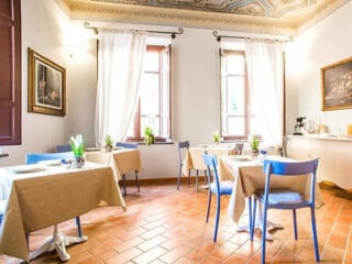 Bright dining room with four tables, blue chairs, potted plants, tile floor, two large windows with white curtains, and framed artwork on the walls.
