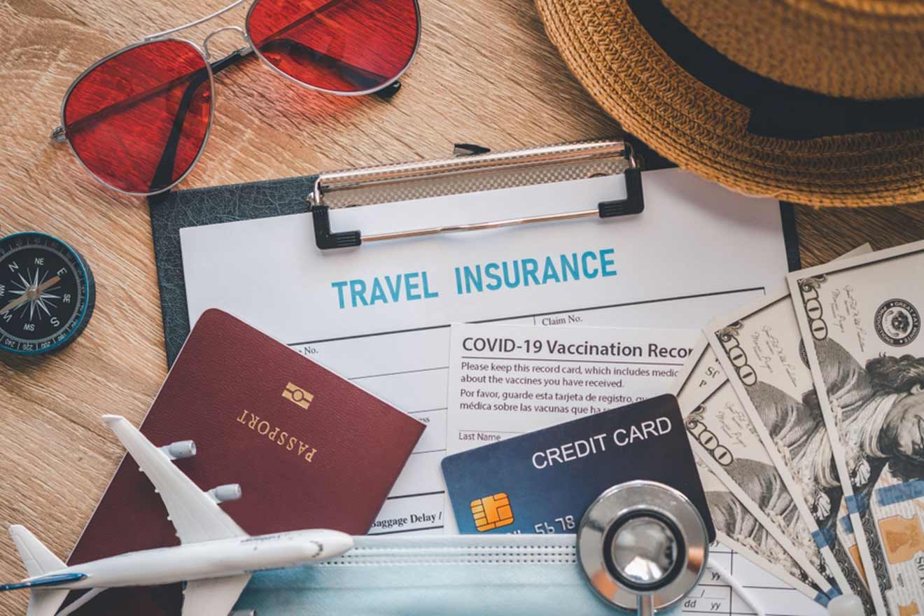 A travel insurance form on a clipboard, a COVID-19 vaccination record card, a passport, a toy airplane, a credit card, US dollar bills, a compass, a hat, and sunglasses on a table.