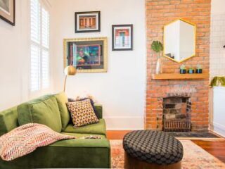 A living room with a green sofa, patterned pillows, a round ottoman, a brick fireplace, framed artwork on the wall, and a gold-framed mirror above the mantel.