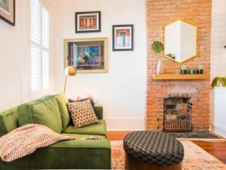 A living room with a green sofa, patterned pillows, a round ottoman, a brick fireplace, framed artwork on the wall, and a gold-framed mirror above the mantel.