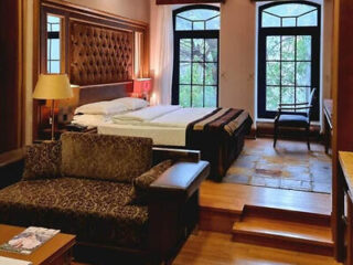A hotel room with a double bed, padded headboard, sofa, desk, lamp, and two large windows overlooking trees.