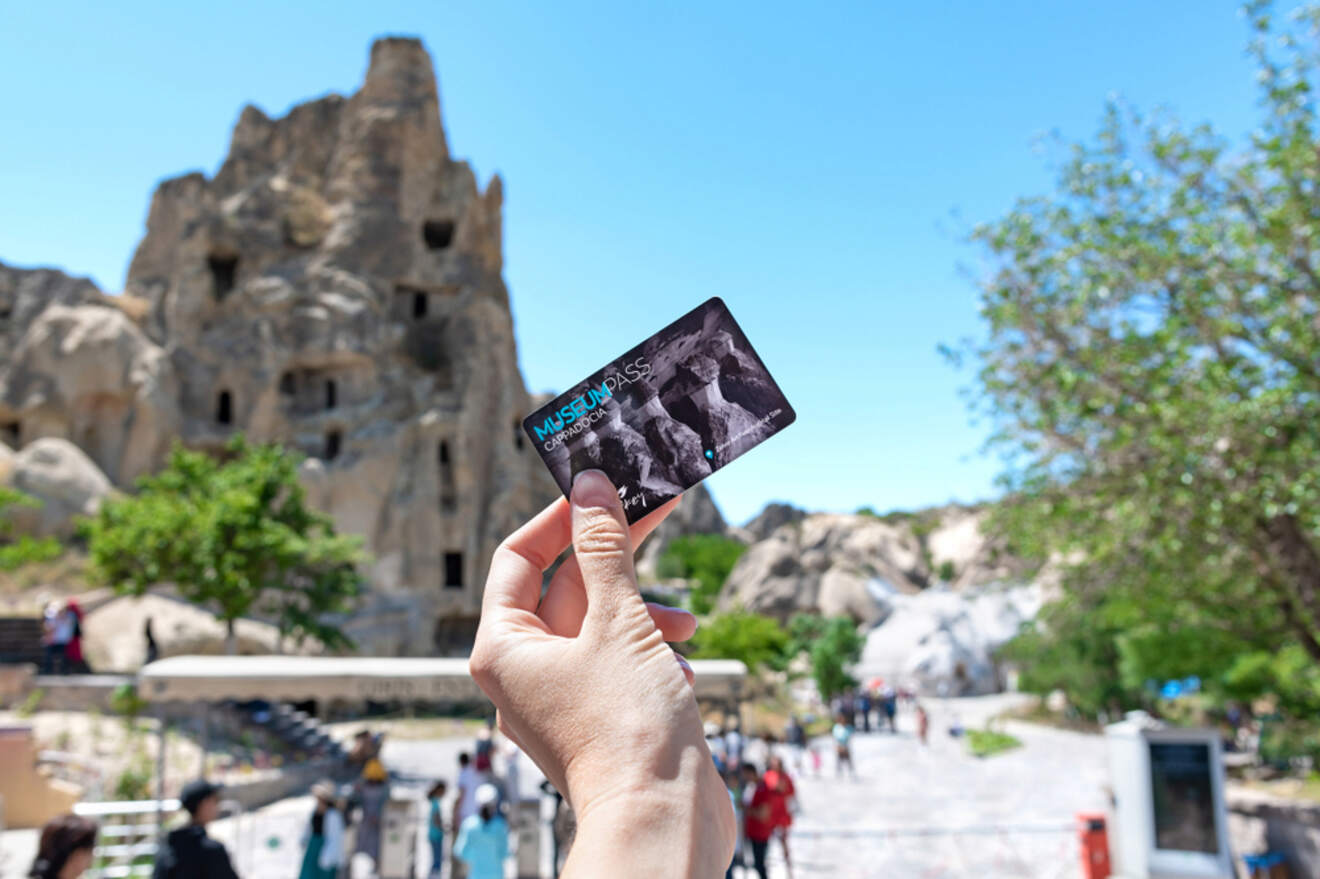 A hand holding a Museum Pass in front of a historic rock formation with people walking around in the background on a sunny day.