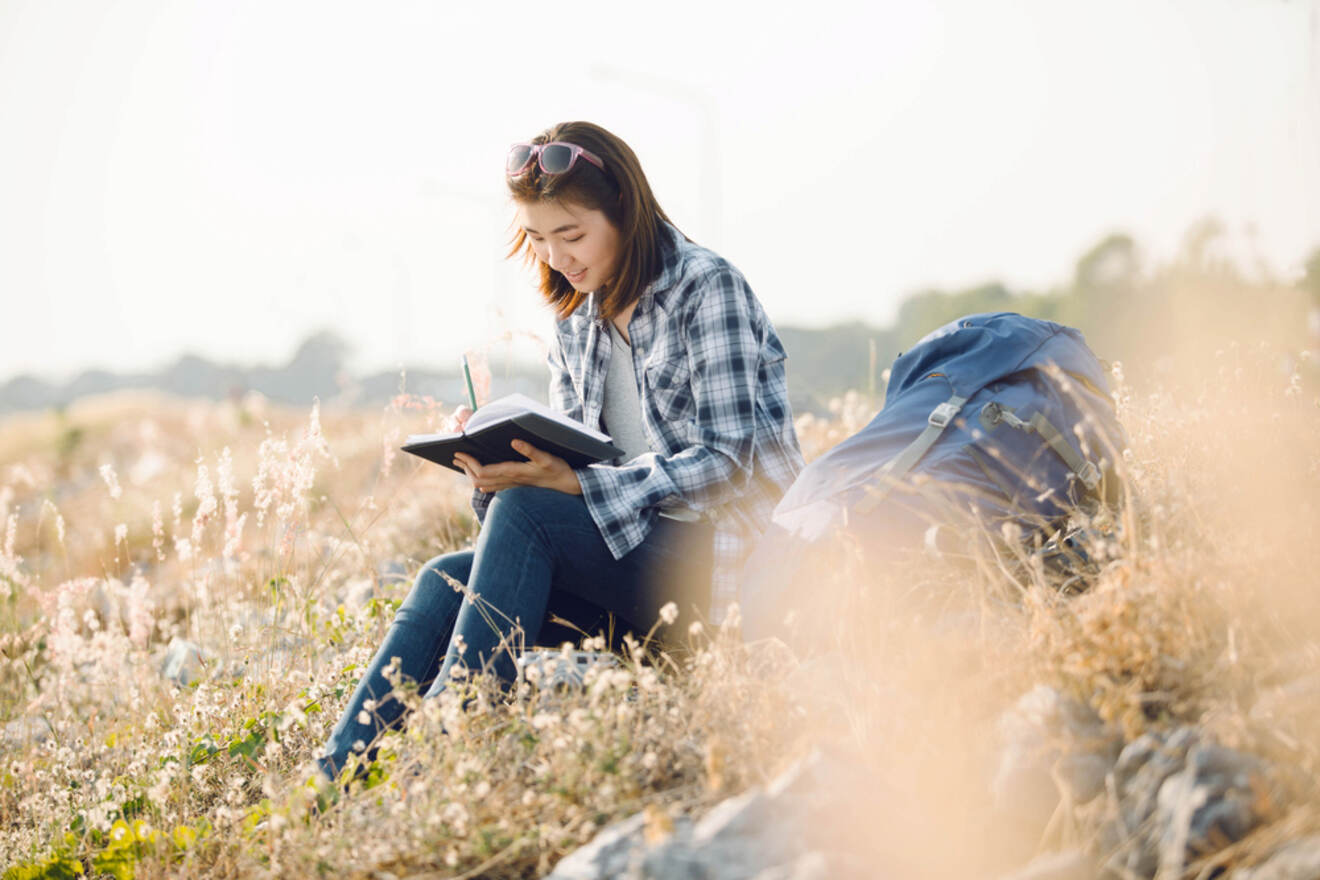 A person sits outdoors on a grassy field, writing in a notebook. A backpack is placed beside them. They are surrounded by dry, tall grass and the sky appears overcast.