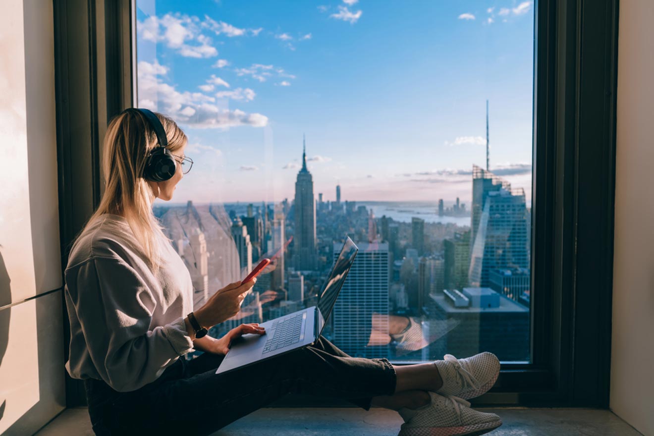 Woman wearing headphones sits by a large window, holding a smartphone and using a laptop. She looks out over a city skyline at sunset.